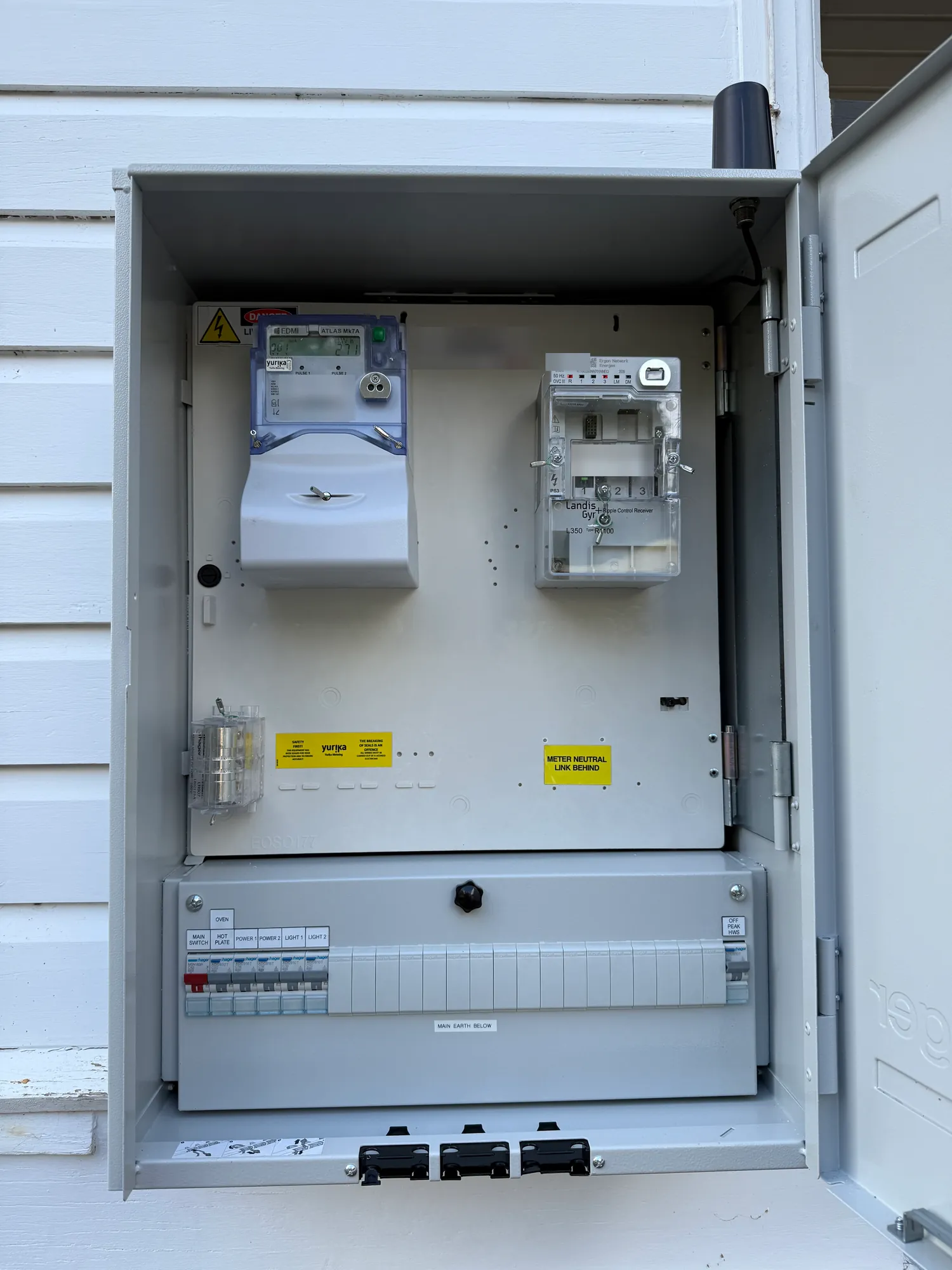 New meter box open on a white weatherboard wall: meters in the top half, a Hager board with printed labels below