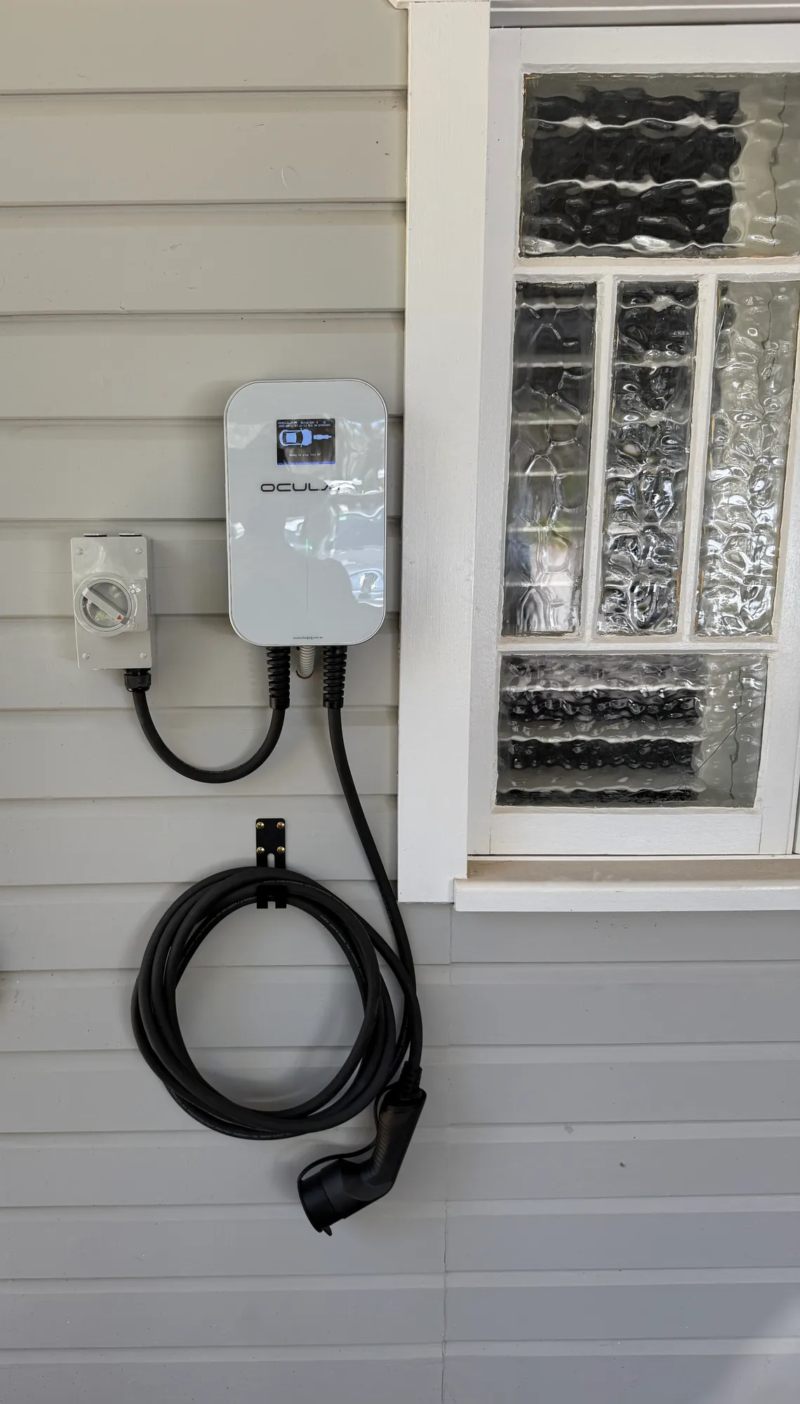 Ocular EV charger on a grey weatherboard wall with an isolator beside it and the charging cable coiled on a wall hook