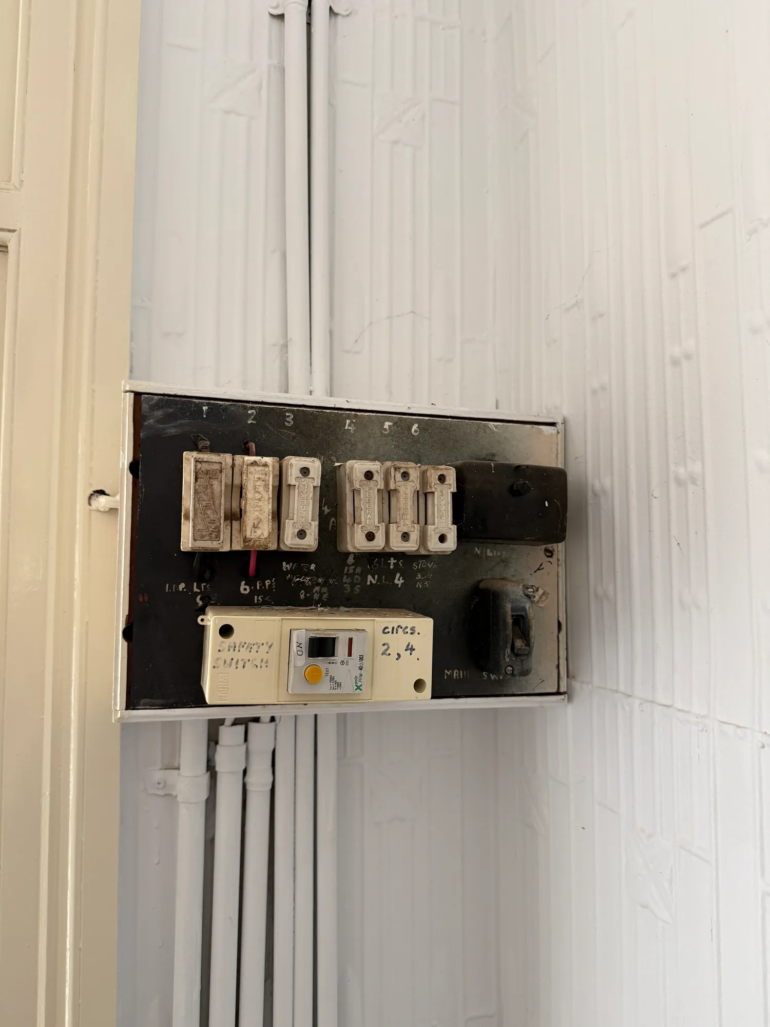 The old setup Glenn found at an older home: rewireable porcelain fuses on a pressed-metal hallway wall, an add-on safety switch that only covered two circuits, and the original dial meters out on the veranda.